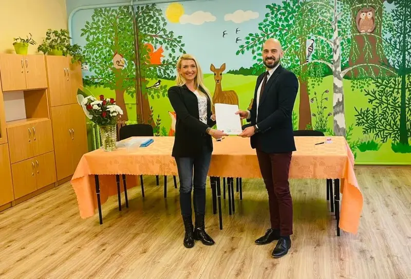 Zdjęcie o orientacji poziomej przedstawia kobietę i mężczyznę stojących w jasnej sali przedszkolnej, którzy wspólnie trzymają biały dokument. Kobieta po lewej stronie ma na sobie czarną marynarkę i ciemne jeansy, natomiast mężczyzna po prawej jest ubrany w granatową marynarkę, białą koszulę i bordowe spodnie. Przed nimi znajduje się stół przykryty brzoskwiniowym obrusem, na którym widać bukiet czerwonych róż oraz notatnik. W tle ściana jest ozdobiona kolorowym malowidłem ściennym z motywami leśnymi, takimi jak drzewa, sarna, wiewiórka i sowa. Po lewej stronie kadru widoczne są wysokie, drewniane szafki, a na podłodze panele o jasnym odcieniu drewna.