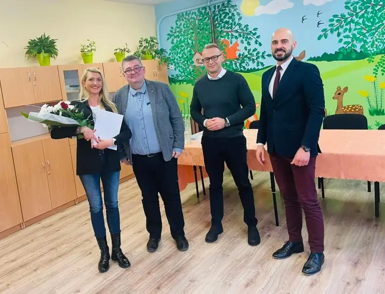 Zdjęcie w orientacji pionowej przedstawia grupę czterech osób stojących w jasnej sali przedszkolnej lub szkolnej. Po lewej stronie znajduje się kobieta w czarnym żakiecie i jeansach, trzymająca bukiet kwiatów oraz białą teczkę, a obok niej stoi trzech mężczyzn w strojach o różnym stopniu formalności – od codziennego po elegancki garnitur. Tło tworzy ściana z kolorowym malowidłem przedstawiającym leśne zwierzęta oraz rząd drewnianych szafek z doniczkami. Na pierwszym planie widać jasną podłogę z paneli, a po prawej stronie znajduje się stół nakryty łososiowym obrusem. Kompozycja zdjęcia jest symetryczna, z postaciami ustawionymi wzdłuż linii horyzontalnej, co dobrze sprawdza się w kontekście dokumentacji oficjalnych spotkań czy wizyt w placówkach edukacyjnych.