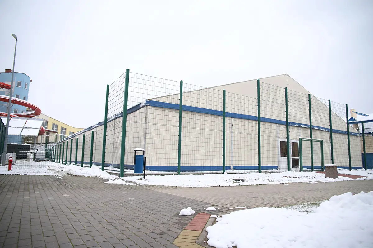 Zdjęcie przedstawia dużą halę namiotową o jasnych ścianach z niebieskimi akcentami, otoczoną wysokim zielonym ogrodzeniem z siatki. Obiekt znajduje się na otwartym terenie pokrytym częściowo śniegiem, z wybrukowaną alejką na pierwszym planie, a całość zachowana jest w proporcjach 3:2. Po lewej stronie widać fragment kolorowej zjeżdżalni wodnej oraz niebieski budynek, co sugeruje lokalizację w sąsiedztwie aquaparku. Niebo jest pochmurne i jasne, co sprawia, że oświetlenie jest rozproszone i równomierne. Przed halą umieszczono niebieski kosz na śmieci oraz charakterystyczny słupek, a całość architektury sprawia wrażenie tymczasowej konstrukcji sportowej lub rekreacyjnej działającej w warunkach zimowych.